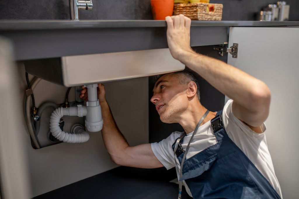 Plumber, repair. Experienced attentive middle-aged man carefully examining bottom of kitchen sink touching pipe with his hand diagnosing problem